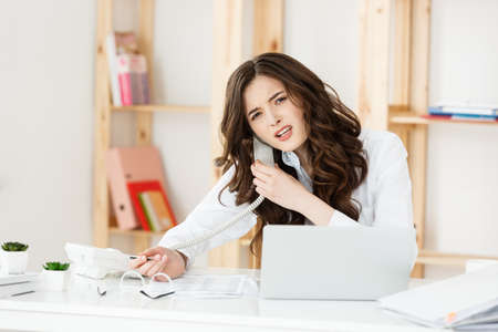 Serious well-dressed saleswoman talking on phone in office behind her desk and laptop computer. Copy spaceの写真素材
