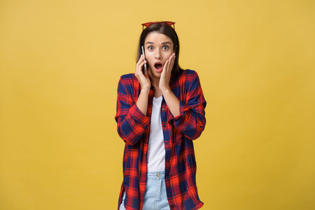 Photo of emotional shocked young pretty woman standing isolated over yellow background using mobile...