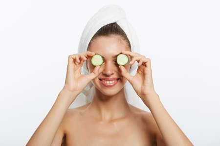 Close up beauty portrait of a happy beautiful half naked woman with a towel wrapped around her head holding cucumber slices at her face isolated over white backgroundの写真素材