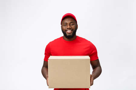 Delivery Concept - Portrait of Happy African American delivery man in red cloth holding a box package. Isolated on Grey studio Background. Copy Spaceの写真素材