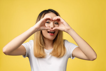 Young Asian woman show heart hand sign isolated on yellow background.