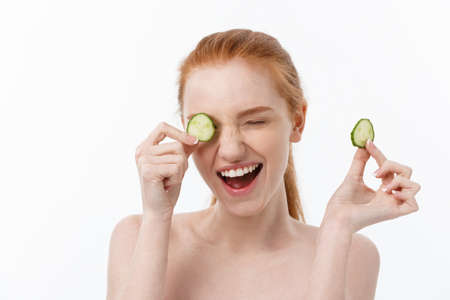 Close up beauty portrait of a smiling beautiful half naked woman holding cucumber slices at her face isolated over white backgroundの写真素材