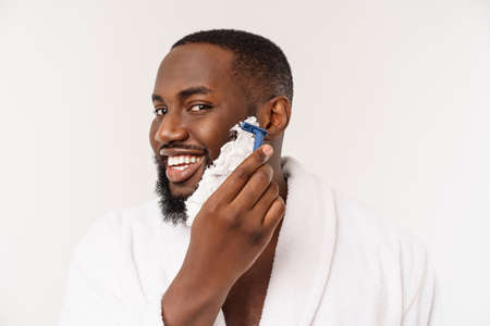 African American man smears shaving cream on face by shaving brush. Male hygiene. Isolated on white background. Studio portraitの写真素材