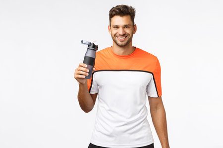 Fitness trainer remind you drink water. Handsome smiling, cheerful sportsman in activewear, finish morning jogging, workout gym practice, grin joyful, holding bottle, standing white backgroundの写真素材