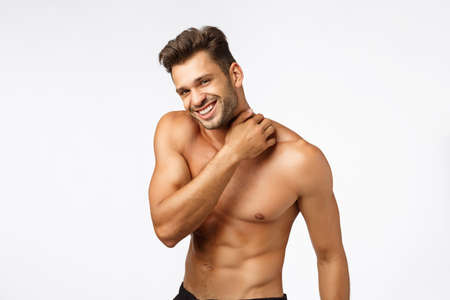 Sport, emotions and beauty concept. Handsome happy young sportsman with six-packs, touching neck and smiling camera with sexy expression, athlete posing naked torso, finish workout, white backgroundの写真素材