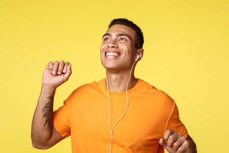 Cheerful carefree man enjoying perfect sunny day during day walk, raising one hand fist pump, dancing joyfully, smiling, enjoy perfect sound in headphones, listen music earphones, yellow backgroundの写真素材