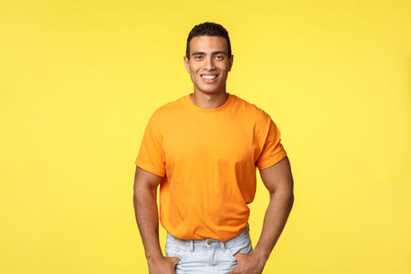 Happy attractive caucasian man in orange t-shirt, hold hands in jeans pockets, smiling upbeat,...