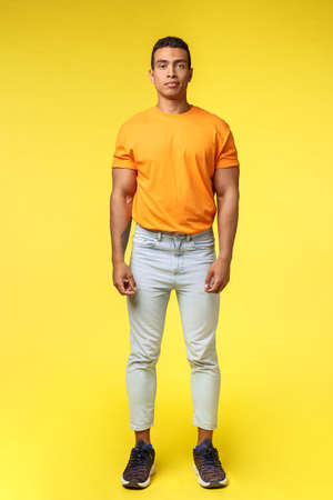 Full-length vertical shot masculine hipster guy, hispanic ethnicity, standing casually yellow background, wear stylish orange t-shirt, white pants, look camera with no expression, slightly smilingの写真素材