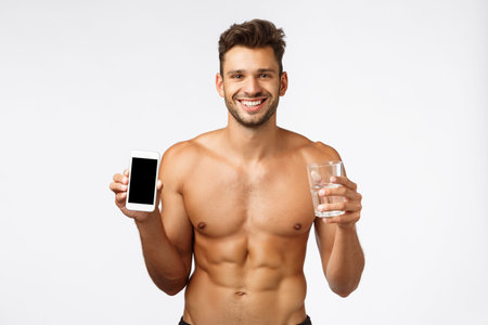 Healthy good-looking shirtless sexy male athlete holding glass water, smartphone, track down how...