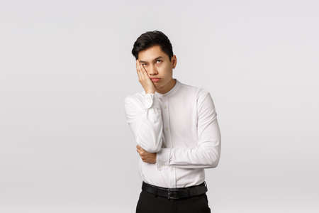 Bored and annoyed young asian businessman dying from boredom and annoyance, roll eyes up, facepalm, lean face on hand and waiting when meeting over, standing irritated white backgroundの写真素材