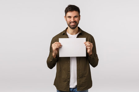 Assertive smiling cheerful bearded caucasian man in coat, holding blank paper near chest and...