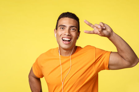 Cheerful, lucky smiling man in orange t-shirt, listen motivational music in headphones, showing...
