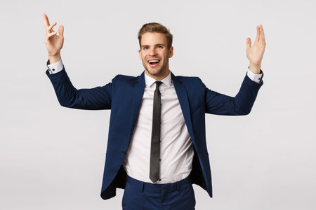 Cheerful lucky and successful male entrepreneur in classic suit, raising hands up delighted, achieve...