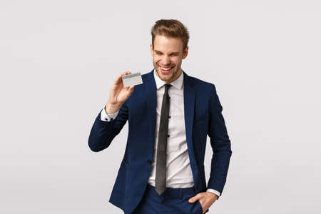 Cheerful good-looking young blond bearded, successful male entrepreneur in classic suit, tie, laughing carefree, showing credit card, paying for purchase, shopping to boost mood, white backgroundの写真素材