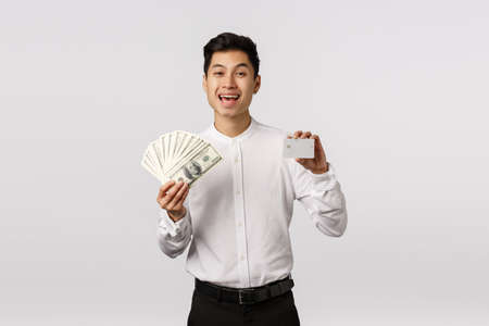 Happy and rich, successful asian guy in formal outfit, holding cash and credit card, laughing and smiling, bragging financial stability, have two variants of payment, picking bank, white backgroundの写真素材