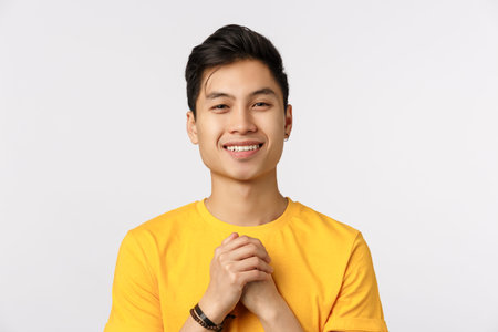 Close-up shot grateful, cute chinese hipster guy in yellow t-shirt, clench hands together over chest...