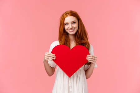 Tenderness, love and romance concept. Romantic charming redhead girlfriend prepared cute gift valentines day, showing red heart card and smiling, express affection and sympathy, pink backgroundの写真素材
