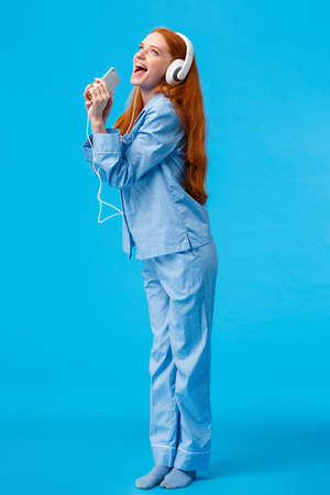 Girl dreaming become star. Vertical full-length portrait charming and carefree redhead teenager in nightwear, donwload karaoke app, listening music in headphones, singing into smartphoneの写真素材