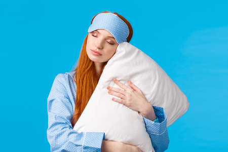 Beauty, female and lifestyle concept. Relaxed and relieved lovely redhead woman in sleep mask and pyjama, close eyes sleepy, having sweet dreams, hugging pillow, standing blue backgroundの写真素材