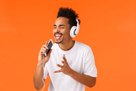 Passionate and carefree african-american guy dreaming become real singer, holding smartphone and...