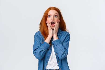 Oh my gosh, amazing. Surprised speechless redhead woman standing starled and shocked, touch face,...