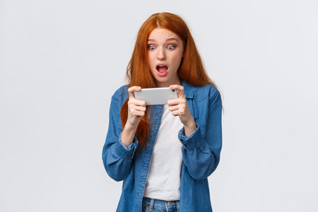 Astonished startled cute redhead woman just won challenge in awesome game, holding smartphone...