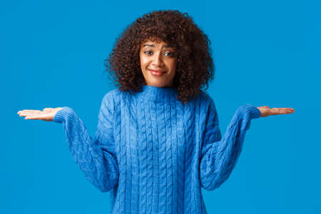 Who cares i dont know. Confused and clueless silly cute smiling african-american female in winter sweater, shrugging with hands spread sideways and smile as have no idea, standing blue backgroundの写真素材