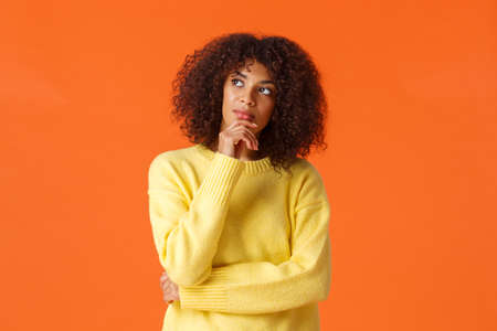 Waist-up portrait unsure, thoughtful young creative african-american female entrepreneur making new ideas, standing orange background, searching inspiration, thinking looking upの写真素材