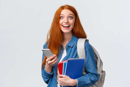 Excited and happy, relieved cute redhead female college student with backpack, notes in notebook and smartphone, looking cheerful camera, texting, messaging friends, white backgroundの写真素材