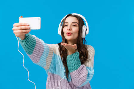 Romance, winter holidays, women concept. Attractive, sensual and flirty young brunette woman in winter sweater, wearing big headphones, holding smartphone and blowing kiss at phoneの写真素材