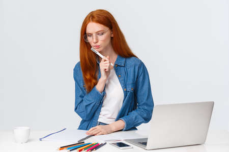 Serious looking focused and determined redhead female working over important project, design for company, work freelance, thinking, pondering as writing, stand near laptop and colored pencilsの写真素材