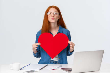Lovely, romantic cute redhead girl folding lips in mwah, send air kiss and holding big red valentines card, showing her love and passion, congratulate girlfriend with anniversary, stand near laptopの写真素材