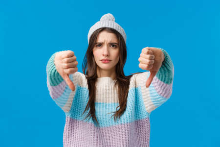 Bad idea, dont do it. Serious-looking upset and angry young woman in winter sweater, hat, looking with judgement, skeptical make thumbs-down gesture, disagree dislike something, give refusalの写真素材