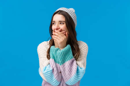 Emotions, fun and happiness concept. Cheerful young caucasian woman in winter sweater, hat, laughing out loud looking aside, laughing close mouth with hands, joking around, standing blue backgroundの写真素材