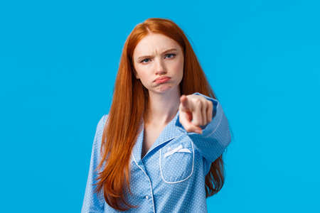 Offended timid and silly redhead teenager, girl in nightwear pointing at camera with blame, shame someone, sulking and frowning angry, accuse person, standing blue background unsatisfiedの写真素材