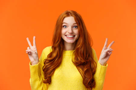 Waist-up portrait lovely and cheerful pretty redhead woman showing peace gestures, smiling camera, travel abroad during winter holidays posing near sightseeing, orange backgroundの写真素材