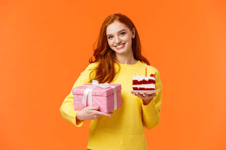 Cute redhead girl coming party with wrapped gift and piece of delicious b-day cake, asking friend blow-out candle and make with celebrating girlfriends birthday, smiling over orange backgroundの写真素材