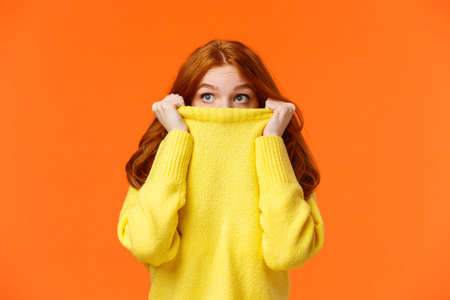 Scared cute teenage redhead girl pulling sweater on face and peeking at upper left corner with frightened eyes, glancing sideways, watching horror movie alone, orange backgroundの写真素材