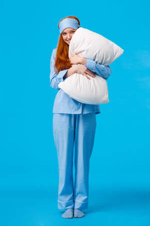 Silly romantic and cute, gentle young redhead coquettish woman in pyjama and sleep mask, hugging pillow peeking at camera with tender lovely smile, standing over blue backgroundの写真素材