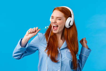 Sing like no one seeing. Carefree and excited joyful redhead female in pyjama, wearing large headphones and singing in toothbrush, close eyes enjoying awesome music, standing blue backgroundの写真素材