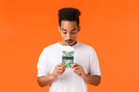 Dreamy and wondered, african-american guy imaging best vacation ever, holding passport with tickets, looking documets amused, standing orange background prepare for flight luxury resortの写真素材