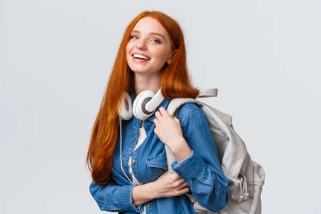 College life, modern lifestyle and education concept. Cheerful good-looking redhead female student with foxy long hair, wearing headphones over neck, backpack, smiling cameraの写真素材