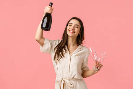 Party, holidays and celebration concept. Cheerful lucky young brunette girl having fun, dancing raising bottle champagne and glass, close eyes and smiling carefree, standing pink backgroundの写真素材