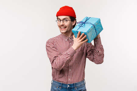 Waist-up portrait happy and excited cute bearded guy trying guess whats inside box, shaking wrapped gift curious what friend give him for b-day, smiling delighted and cheerful, stand white backgroundの写真素材