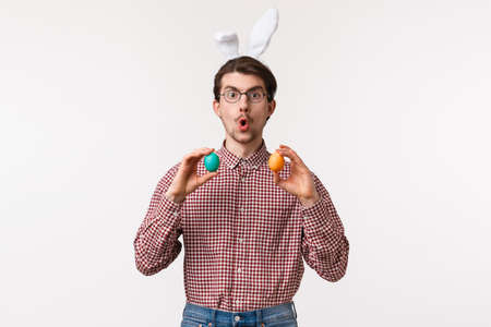 Traditions, religious holidays, celebration concept. Amused enthusiastic handsome young man suggest girlfriend play Easter game on family gathering party, hold two painted eggs, wear rabbit earsの写真素材
