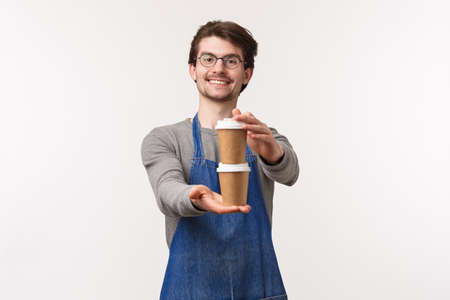 Portrait of friendly cheerful young male employee in apron, working coffee-shop giving customer his order two cups of drink, prepared cappuccino and smiling as saying enjoy your drinkの写真素材