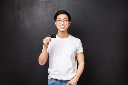Bank, finance and payment concept. Portrait of satisfied charismatic asian guy in t-shirt and glasses, recommend put money on deposit, pleased with banking service, hold credit card and smilingの写真素材
