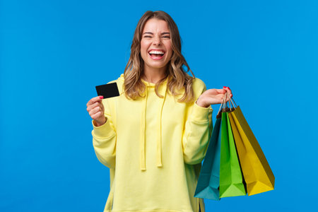 Carefree emotive, smiling happy pretty blond girl using credit card to waste some money in mall,...