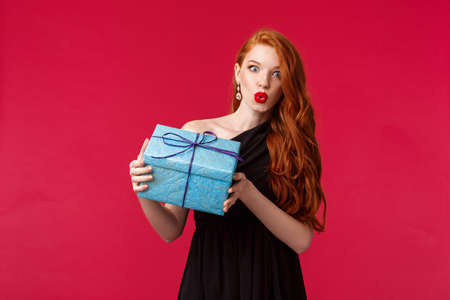 I wonder whats inside. Curious stunning young redhead woman in elegant black dress, shaking box with gift, folding lips amazed and intrigued look camera, receive present, red backgroundの写真素材