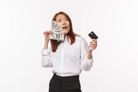 Success, women and career concept. Satisfied pleased and boastful young asian female enjoying being rich, holding credit card and brush cheeks with dollars, have lots money, white backgroundの写真素材